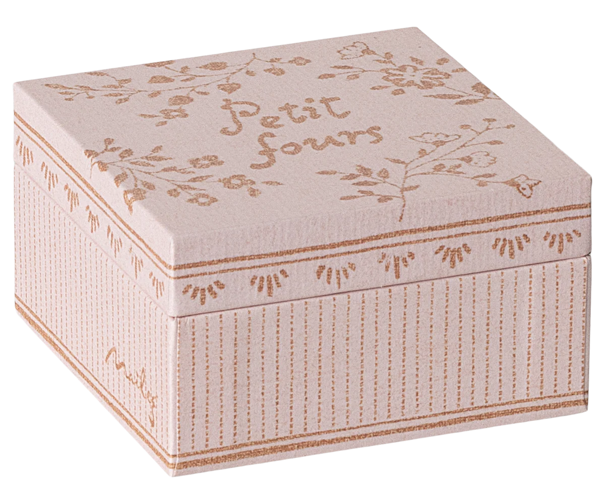 A square, light pink Maileg Cakes Display box with delicate floral patterns and "Petit fours" in brown handwritten style on the lid. Perfect for dollhouse mini cake sets, it features decorative lines and tiny flowers along the sides. Brand: Maileg.