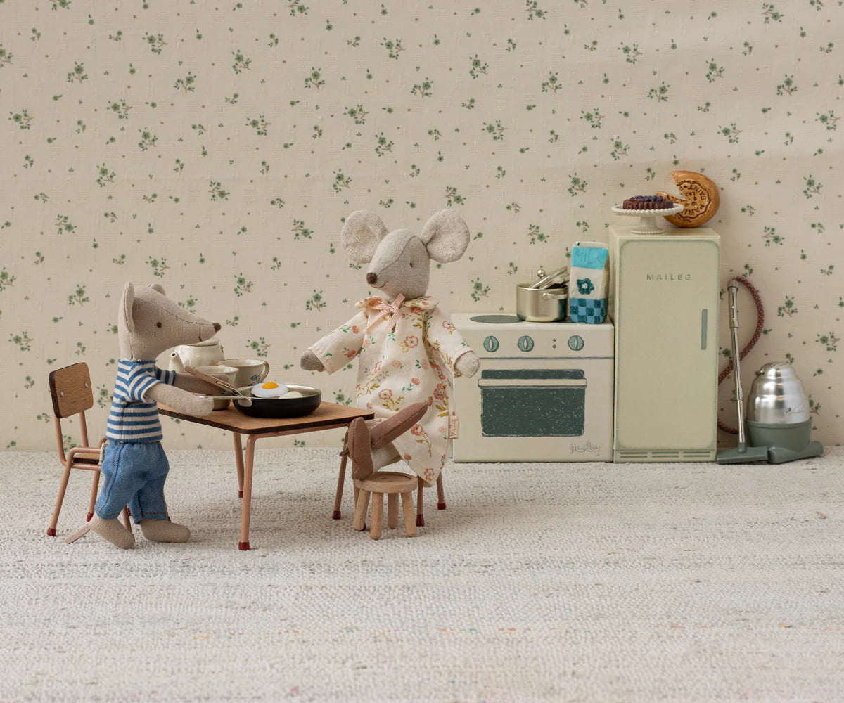 Two plush mice enjoy breakfast in a miniature Mouse hole Farmhouse kitchen. One mouse sits at the Maileg Table and Chairs by Maileg, while the other stands nearby in pajamas amid vintage appliances and tiny plates of food.
