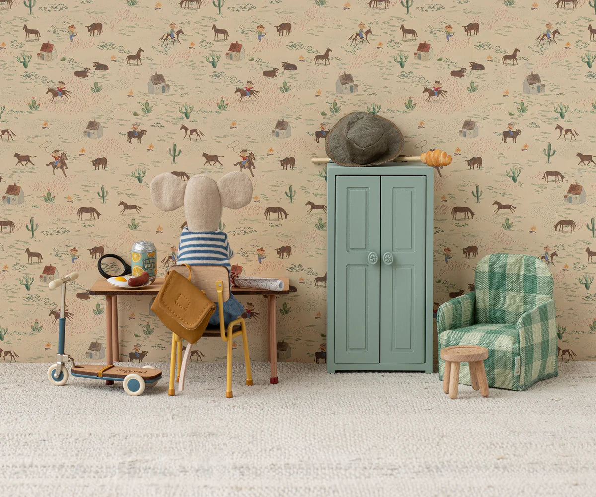 A Maileg fabric mouse doll sits at a small desk with a vintage seat and scooter nearby, facing western-themed wallpaper. To the right is a Maileg Yellow Chair, plus a blue cabinet with a hat and bread on top.