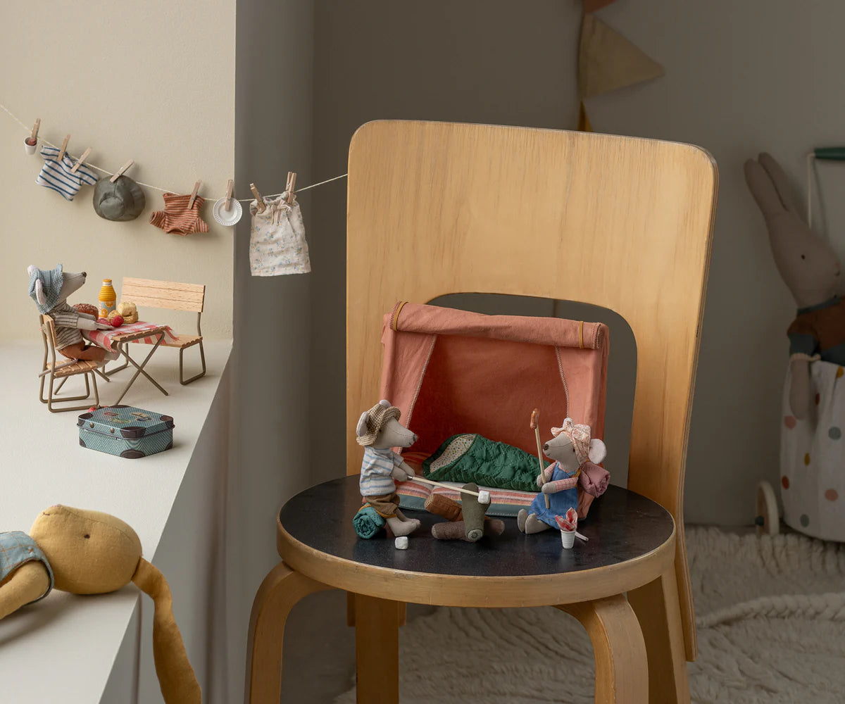 Three Maileg toy mice play: one lounges at a picnic table by the window, while two gather near the Maileg Happy Camper Mouse Tent on a wooden chair. Tiny clothes hanging on a string complete their cozy outdoor scene.