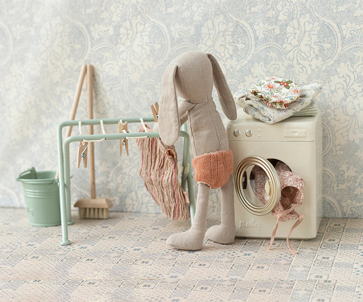 A Maileg fabric bunny doll in orange shorts stands by the Maileg Drying Rack with pegs and a toy washing machine, hanging tiny clothes amid folded fabrics and miniature laundry items against patterned wallpaper.