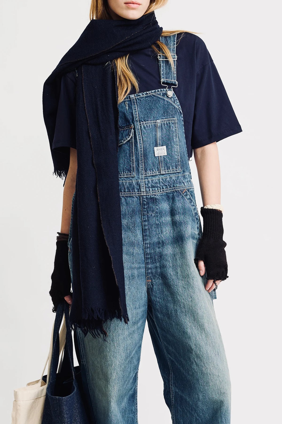 A person in Denimist SLOUCH BIB OVERALL, with a utility pocket, navy tee, dark scarf, black fingerless gloves, and holding a tote bag stands against a plain light background.