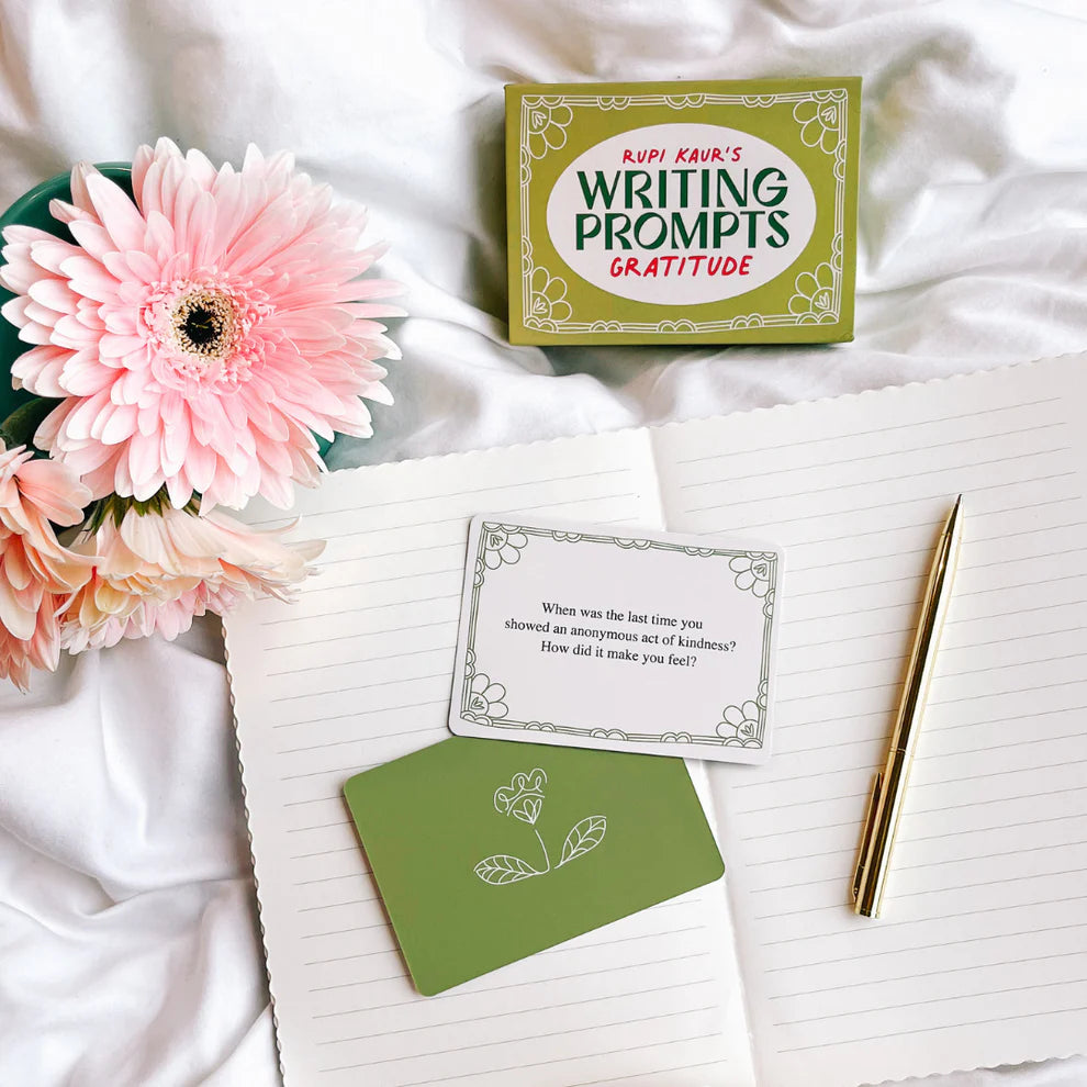 A gold pen lies on an open notebook beside a pink flower and Rupi Kaur's Writing Prompts Gratitude cards by Simon & Schuster Inc. One card asks about a recent act of kindness and its impact.