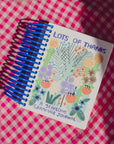 A spiral-bound Faire Gratitude Journal with a colorful floral cover, made in the USA, rests on pink and white checkered fabric.