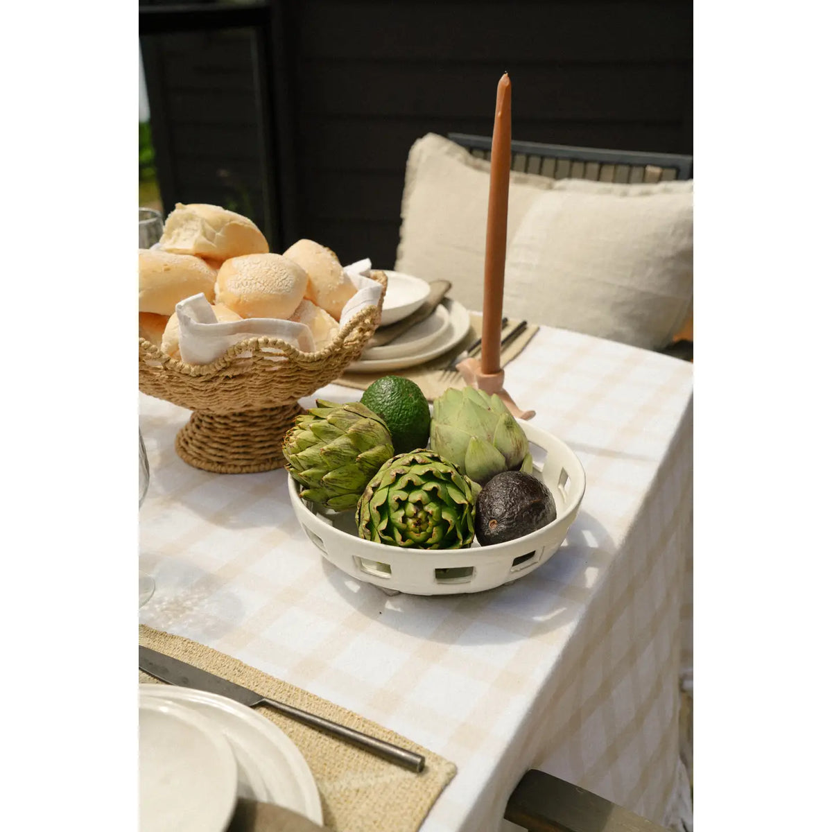 A table set for a meal with a white tablecloth features the Indaba Cypress Fruit Bowl L filled with artichokes and avocados, alongside a basket of bread rolls, beige candle, and cushioned seating—an ideal kitchen accessory for any countertop.