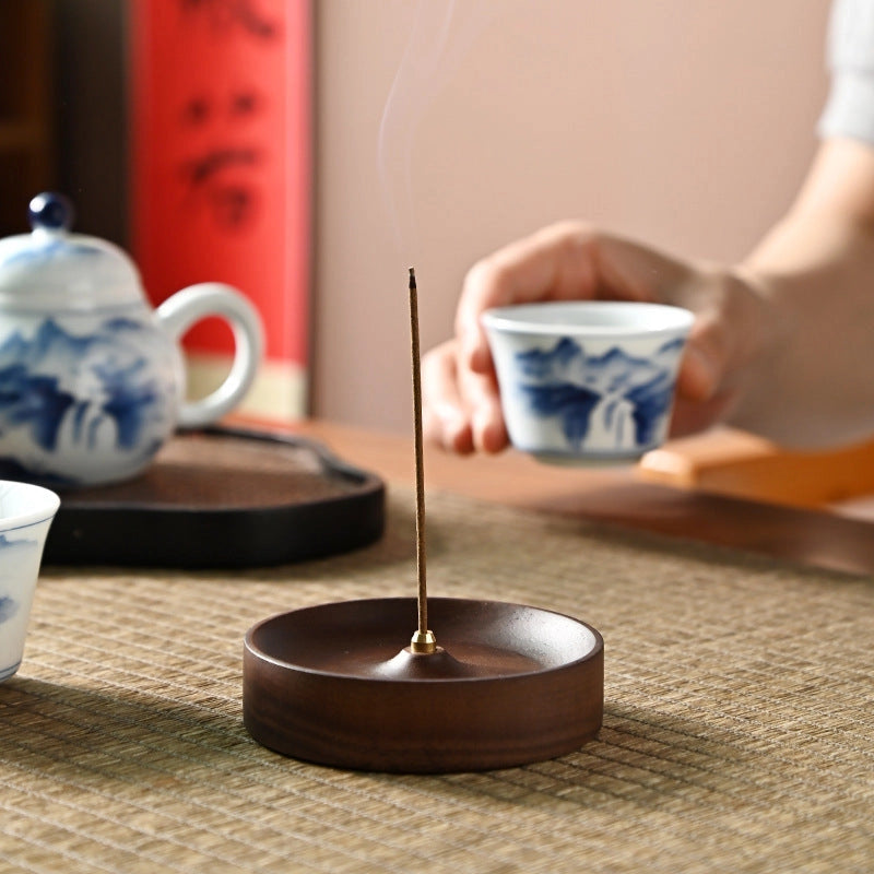 A lit incense stick rests in the Faire Walnut Wood Incense Burner on a woven mat, while a hand holds a blue-and-white teacup. Handcrafted ceramic plates with rustic glaze complete this serene, artisan-inspired tea scene.