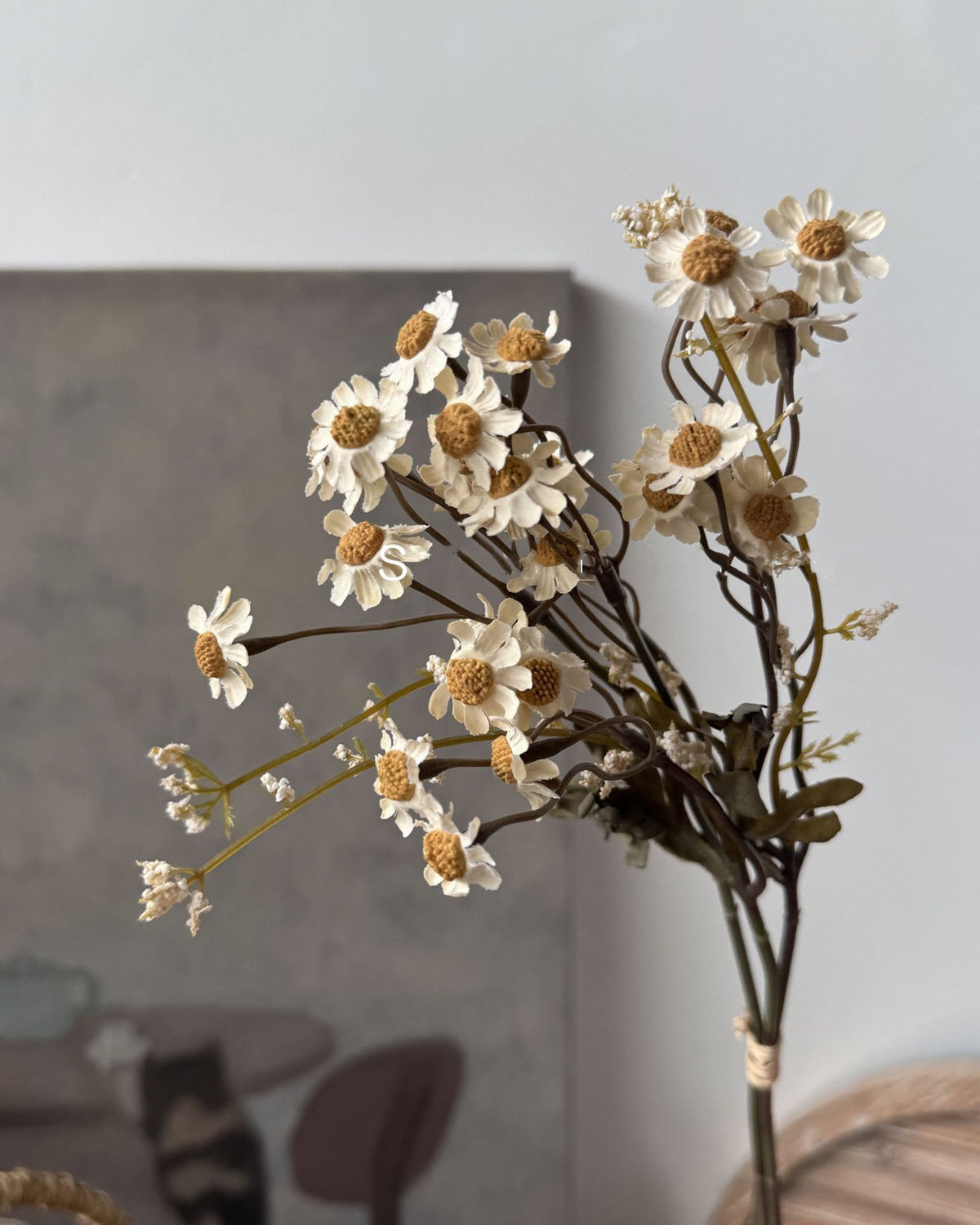 The Faire 13'' Faux Daisies Floral Bunch features small white and yellow daisies with a dried look, set in focus against a softly blurred, neutral background with subtle shapes and hints of furniture.