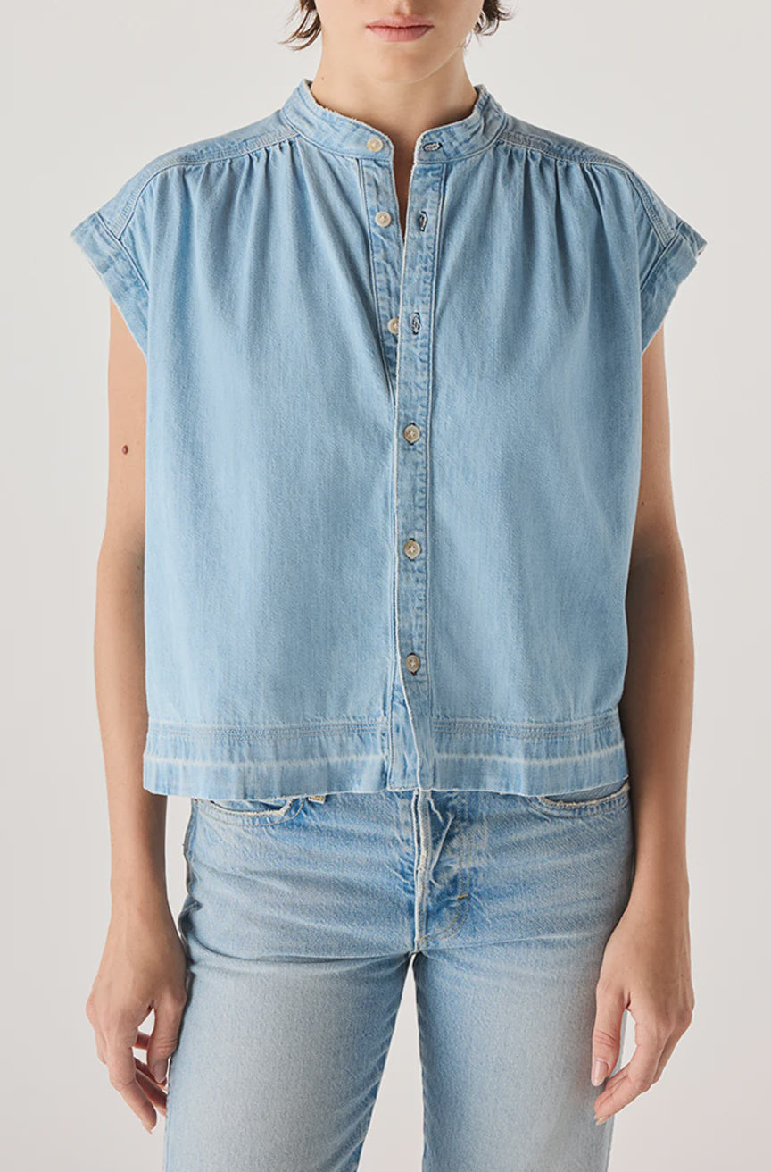 A person wears the Tessa Top by AMO Denim—a light blue, short-sleeved, button-up denim shirt with mandarin collar and relaxed fit, made in Los Angeles from 100% cotton—paired with light blue jeans against a plain background.