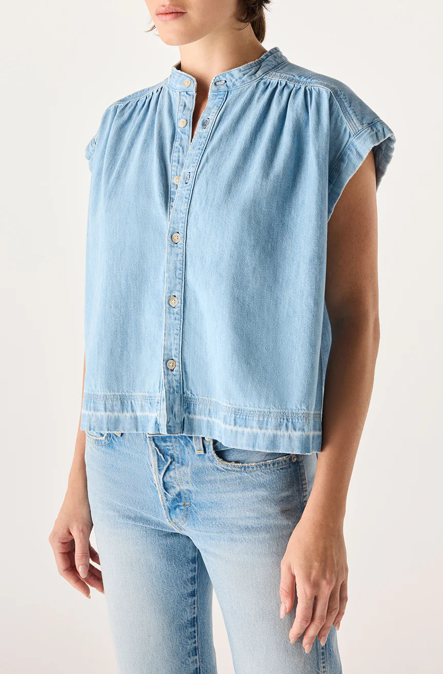 Someone wearing the AMO Denim Tessa Top, a light blue sleeveless button-up denim shirt with a high collar, paired with relaxed fit light blue jeans, made in Los Angeles, stands against a plain background.