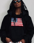 Distressed American flag sweater by Denimist worn under a black hooded jacket.