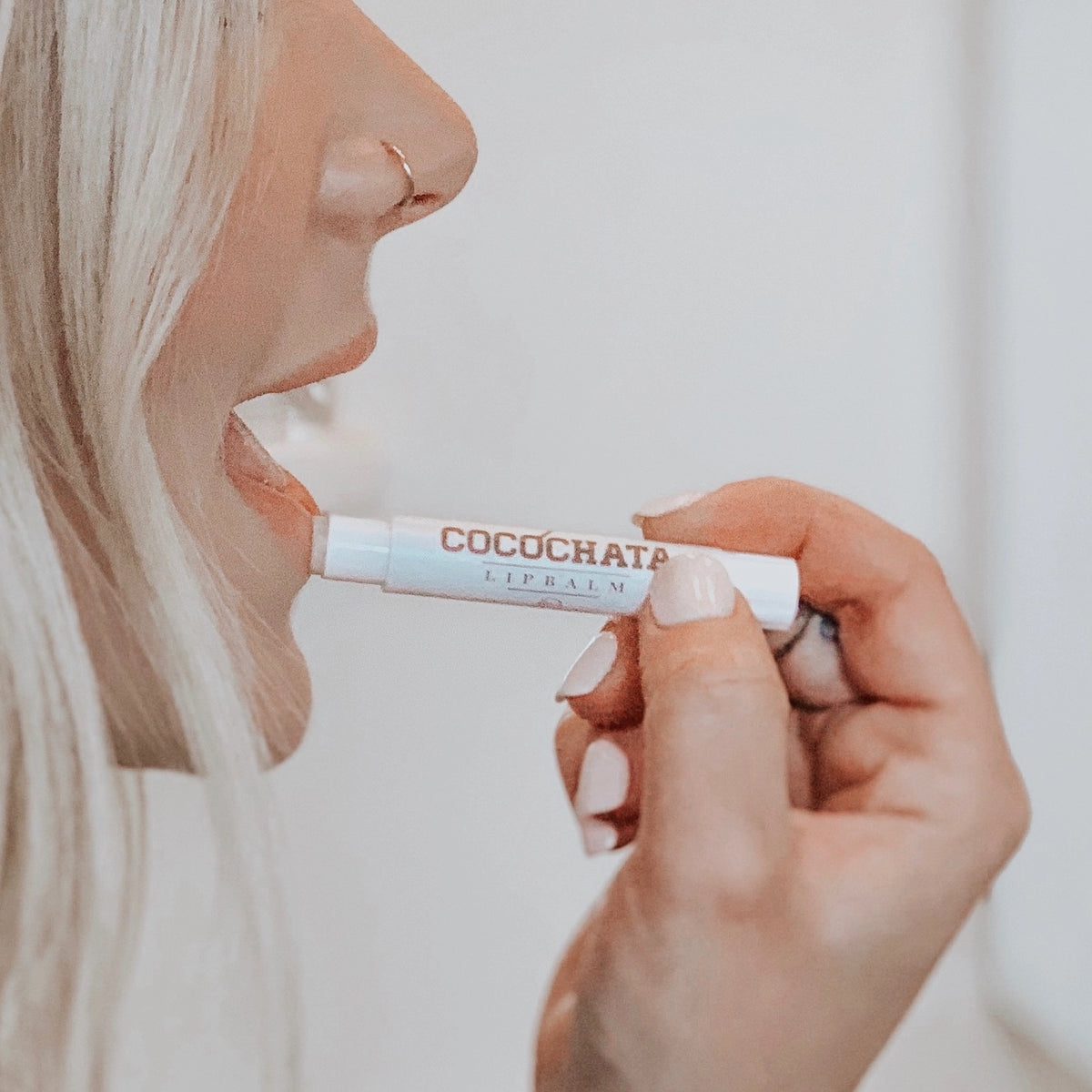 With neatly manicured fingers, a light-haired person applies Faire's Cocochata Lip Balm, made with organic coconut oil, by holding the tube close to their lips.
