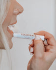With neatly manicured fingers, a light-haired person applies Faire's Cocochata Lip Balm, made with organic coconut oil, by holding the tube close to their lips.