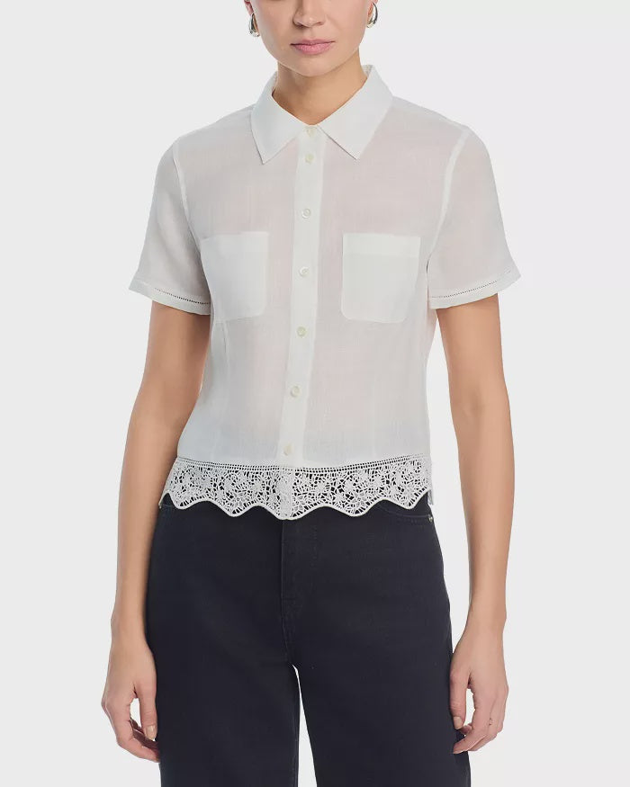 A woman wears FRAME’s Embroidered Seaside Shirt, a short-sleeve sheer white button-down blouse with a collar, two chest pockets, and scalloped lace edges, styled with dark high-waisted pants.