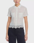 A woman wears FRAME’s Embroidered Seaside Shirt, a short-sleeve sheer white button-down blouse with a collar, two chest pockets, and scalloped lace edges, styled with dark high-waisted pants.