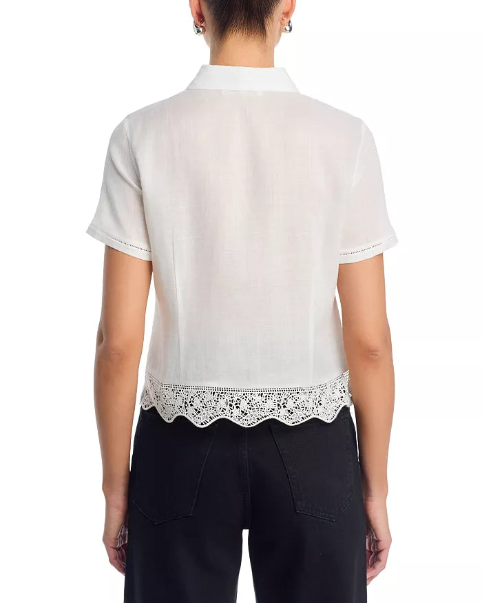 A person wearing the FRAME Embroidered Seaside Shirt—a short-sleeve, white button-down with a scalloped, eyelet lace hem—and black pants, shown from the back.
