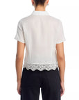 A person wearing the FRAME Embroidered Seaside Shirt—a short-sleeve, white button-down with a scalloped, eyelet lace hem—and black pants, shown from the back.