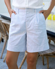 Wearing Frank & Eileen’s Waterford Short in light blue and white stripes with a white shirt, a person stands with hands in pockets by a table and wicker chairs on the patio.