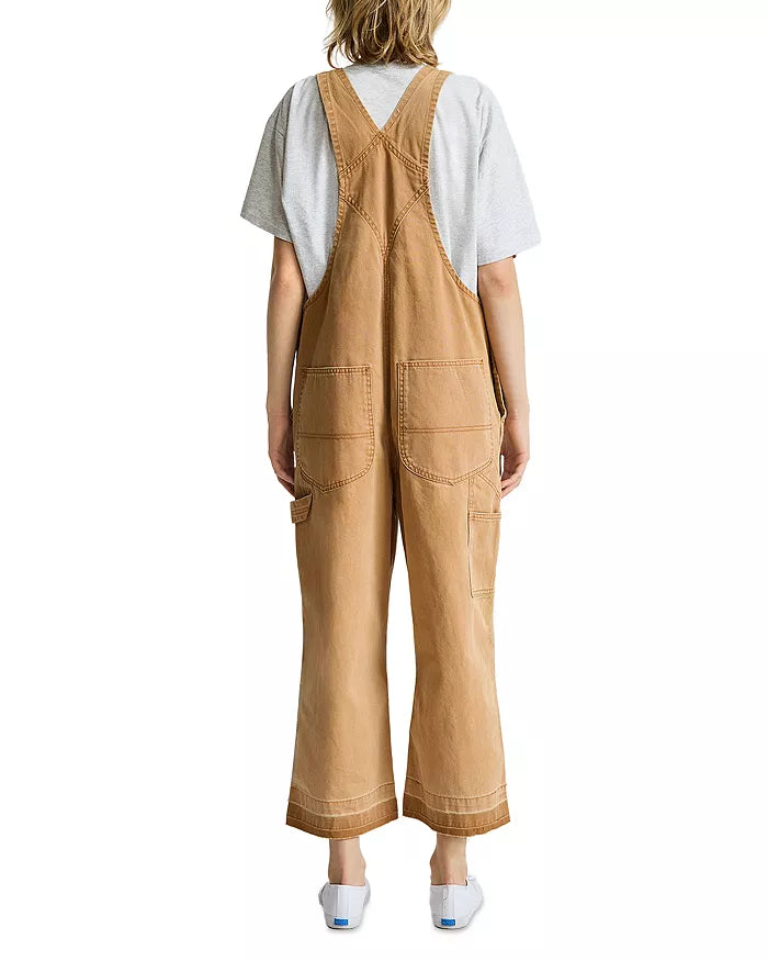 A person with shoulder-length, wavy hair stands with their back to the camera, wearing Denimist’s RELAXED OVERALL in tan—a 100% cotton style featuring adjustable buckle straps, large back pockets, and a relaxed-leg silhouette—layered over a light gray t-shirt and paired with white sneakers.