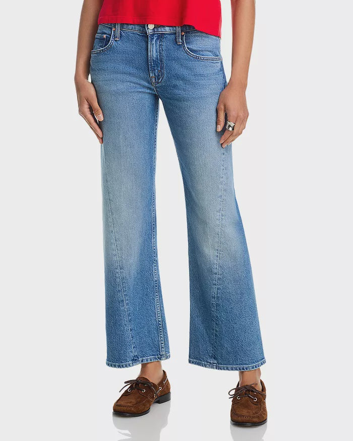 The lower half of a person stands against a white background, wearing Mother Lil Bookie Twist Skimp mid rise flare-leg jeans and brown moccasin-style shoes with a red top.