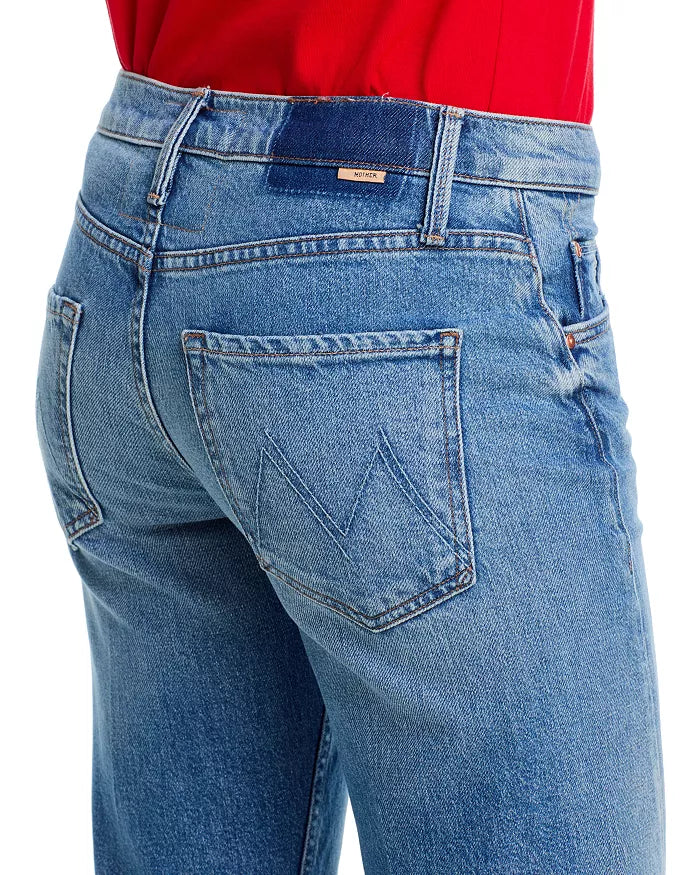 A person is shown from the back wearing Mother’s Lil Bookie Twist Skimp blue denim mid rise jeans with a five-pocket design, stitched "M" on the back pocket, belt loops, and seam details, paired with a red top.
