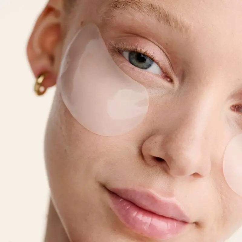 A close-up of a light-skinned person with blue eyes, gold hoop earrings, and a subtle smile wearing Faire Dew Cloud Eye Mask—translucent under-eye gel patches—on both cheeks against a neutral background.