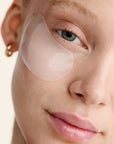 A close-up of a light-skinned person with blue eyes, gold hoop earrings, and a subtle smile wearing Faire Dew Cloud Eye Mask—translucent under-eye gel patches—on both cheeks against a neutral background.