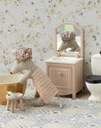 The Maileg Wellness Mouse / Big Sister by Maileg stands in a cute playset bathroom with a sink, mirror, toilet, and stool holding a toothbrush. She wears a towel and shower cap as she leans over the bowl on the patterned floor.