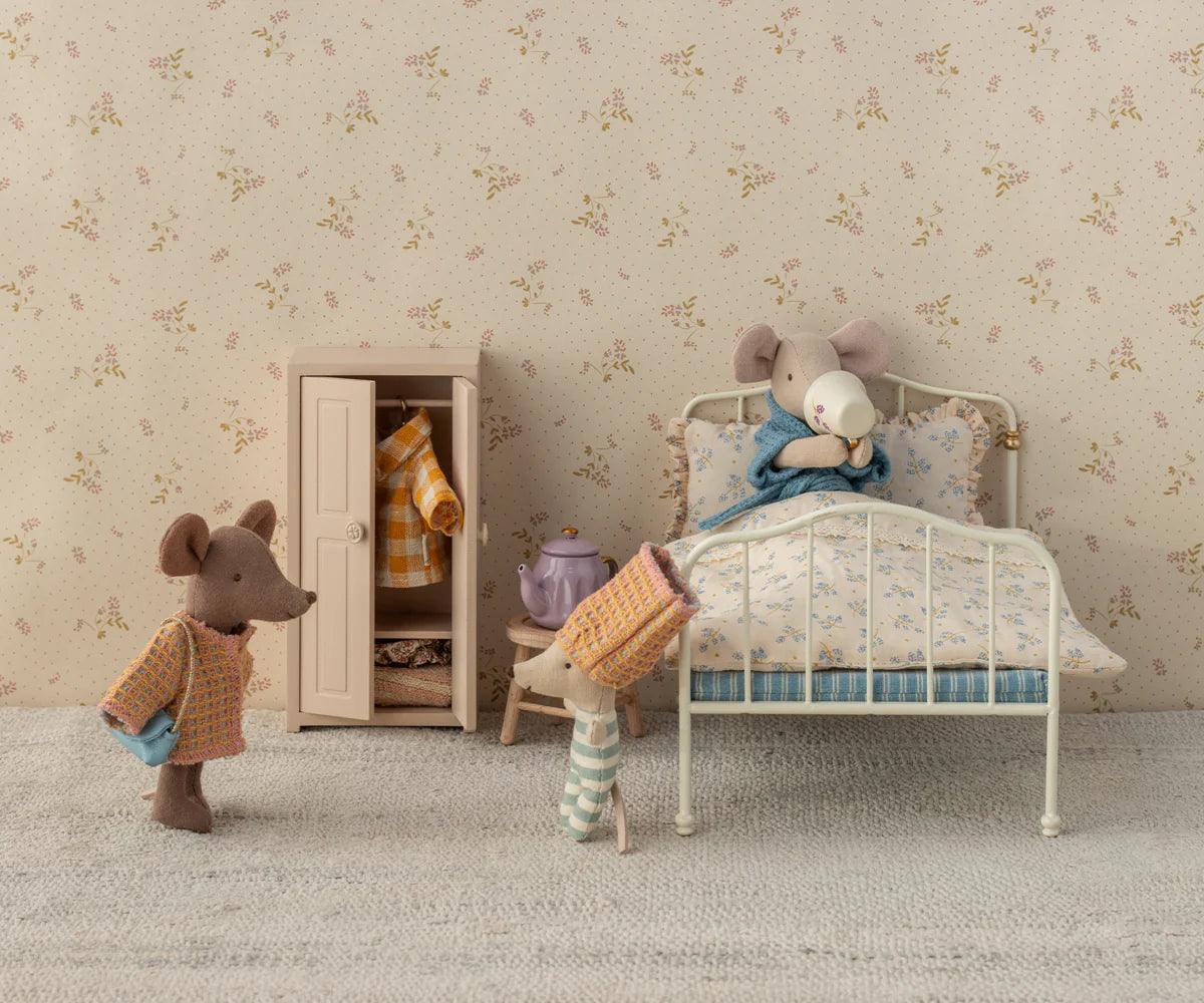 Three Maileg Mum Suit mice relax in a vintage-style bedroom by Maileg: one sits in bed sipping from a mug, one stands near a wardrobe with tiny outfits, and another wears pajamas and a hat. A teapot rests on the bedside table.