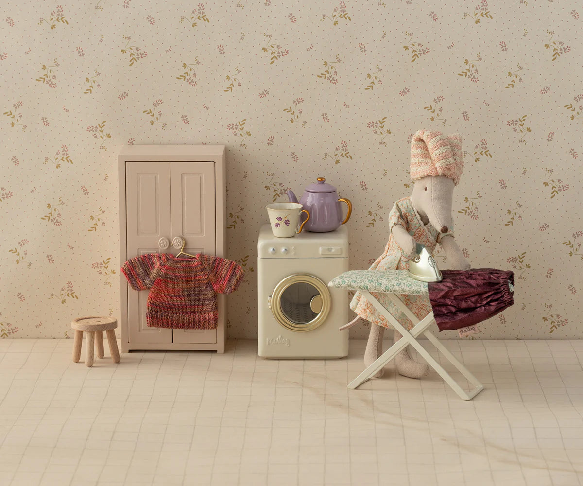 The Maileg Skirt & Knitted Blouse mouse by Maileg, dressed in a skirt and blouse, irons clothes on a dollhouse ironing board next to a washing machine, cupboard, stool, and teapot in a charming laundry room scene.