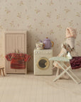 The Maileg Skirt & Knitted Blouse mouse by Maileg, dressed in a skirt and blouse, irons clothes on a dollhouse ironing board next to a washing machine, cupboard, stool, and teapot in a charming laundry room scene.