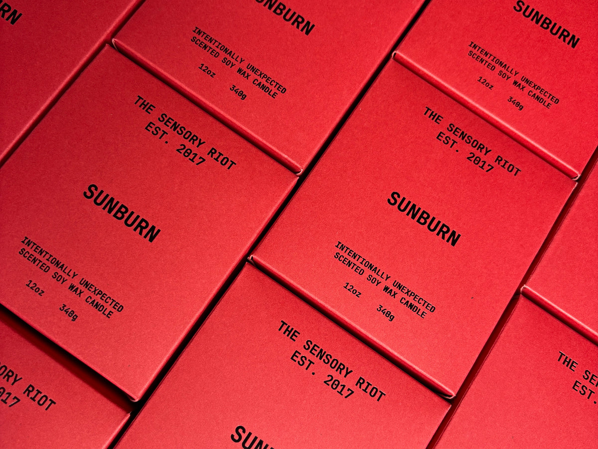 Several red rectangular boxes labeled "SUNBURN 12 oz candle" by Faire are arranged diagonally, showcasing a bold, non-toxic, vegan candle collection.