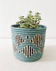 A succulent sits in Faire's Blue Storage Plant Basket: Mifuti, a handmade Rwanda woven planter with teal, black, and cream geometric patterns, displayed on a light surface against a plain white background.