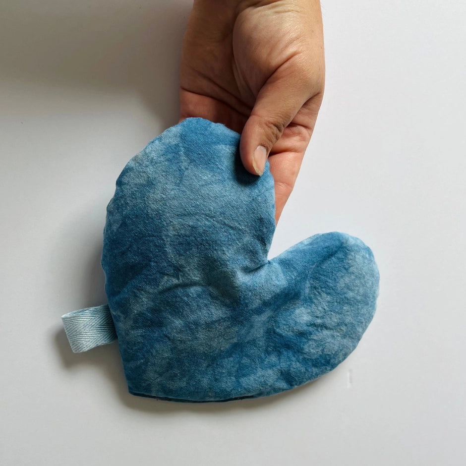 A hand holds the Faire Heart Eye Pillow—a soft, blue, heart-shaped cotton pillow with a fabric loop—perfect for anxiety relief, shown against a plain white background.