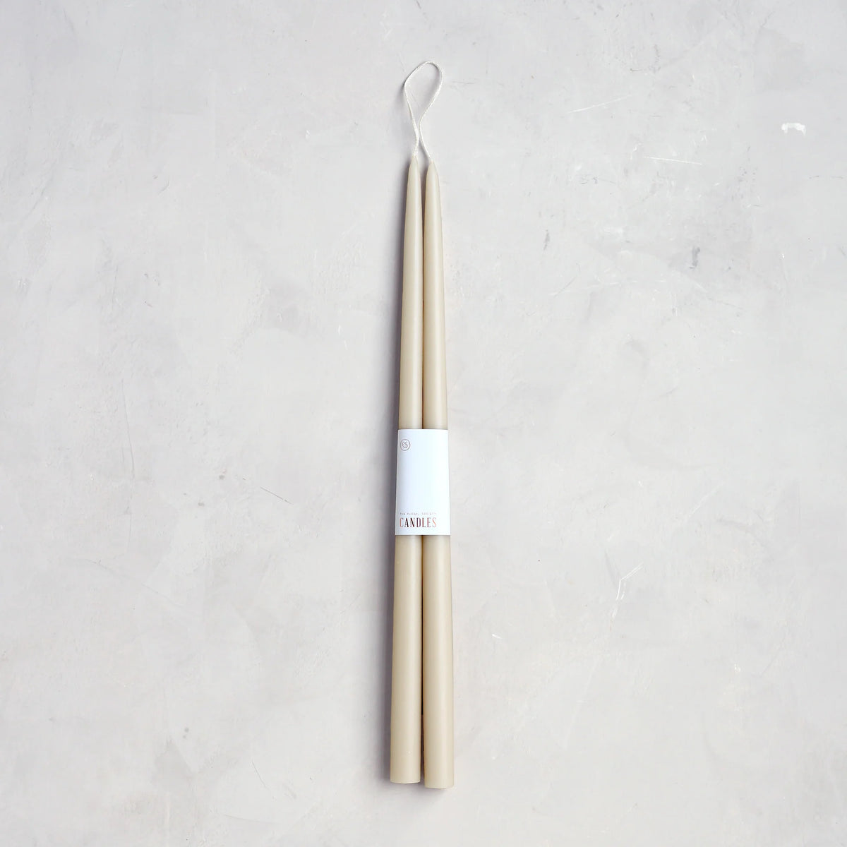 The Floral Society 18" Taper Candle - Pair in beige features a classic design with a single looped wick for hanging. Expertly crafted, they come bundled with a white paper label and rest on a light grey textured surface.