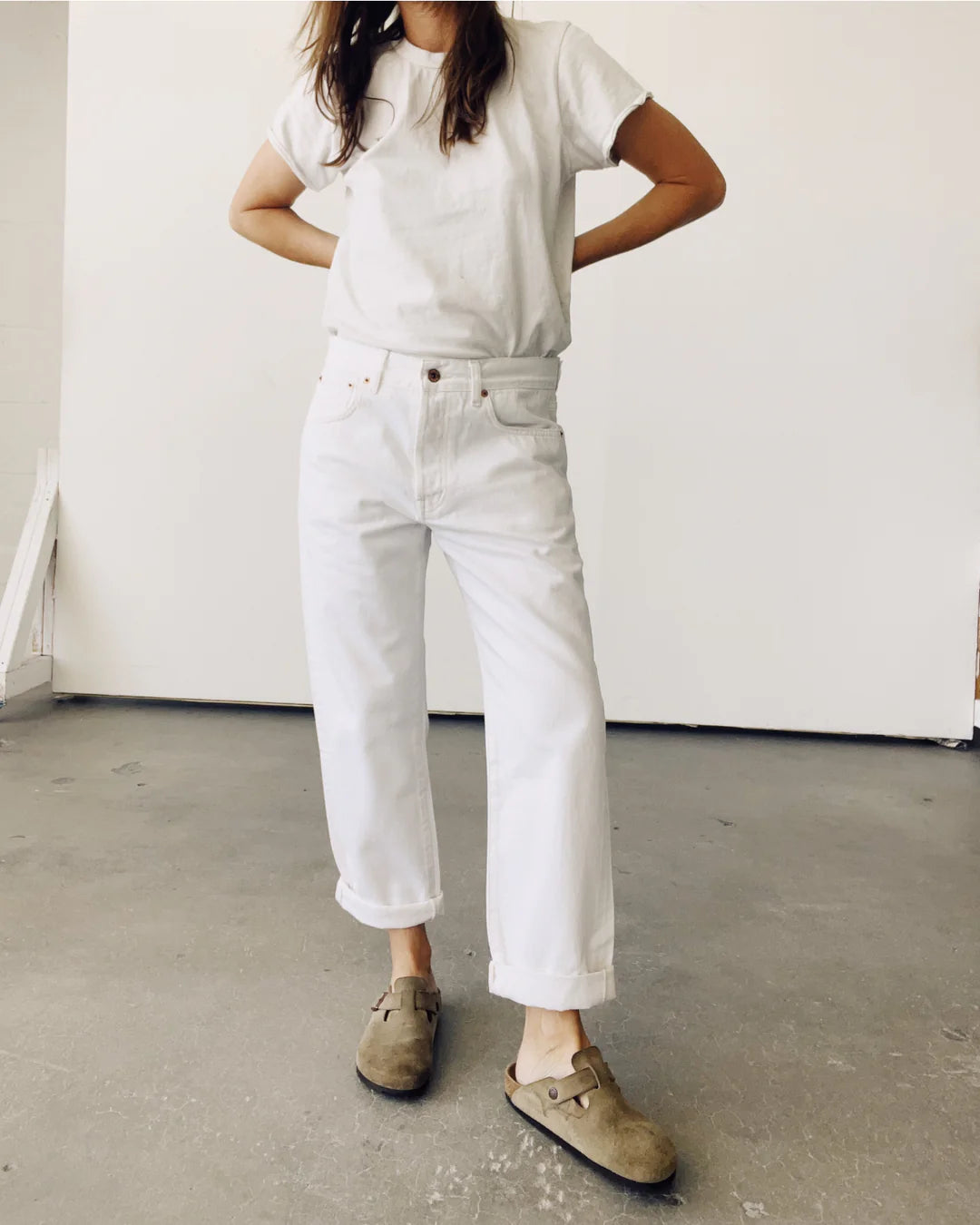 A person stands with hands on hips, wearing a white t-shirt, Hey Gang's "The Loose Crop" jeans made of 100% cotton denim, and brown slip-on shoes. The scene features a plain white wall and concrete floor, with the person's face out of view.
