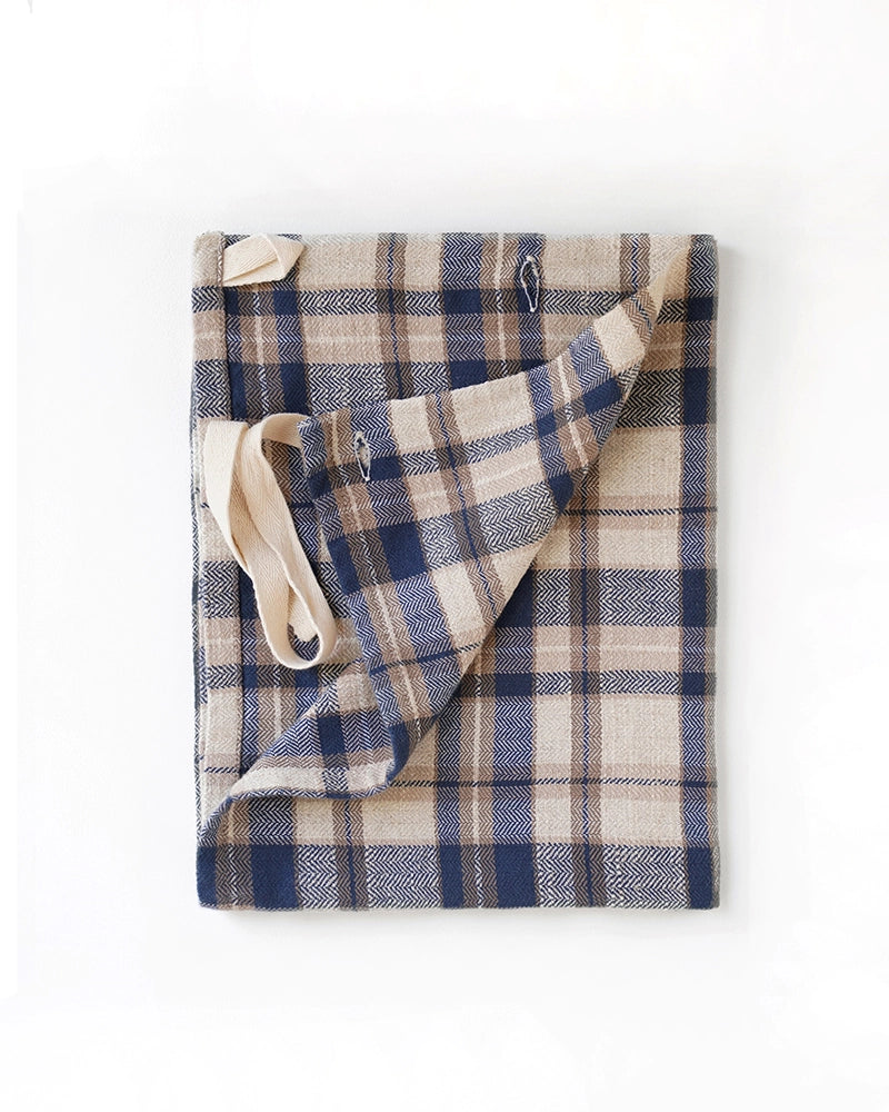 The Faire Cook's Cloth, a beige and navy blue plaid apron with cream straps, made from a cotton-linen blend, is shown folded on a white background.