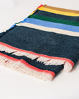 The Faire Multicolor Cabin Stripe Throw Blanket features horizontal yellow, green, blue, cream, and red stripes on a dark denim background with white fringe—ideal for rustic home decor or as a cozy cabin throw.