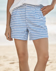 A person wears Frank & Eileen’s Waterford Short in blue and white gingham with a wide-leg fit and a white top, standing on a sandy beach with the ocean blurred in the background. Only the lower torso and upper legs are shown.