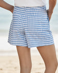 A person wearing Frank & Eileen Waterford Shorts in blue and white gingham with a white top stands on a beach, the ocean blurred in the background.
