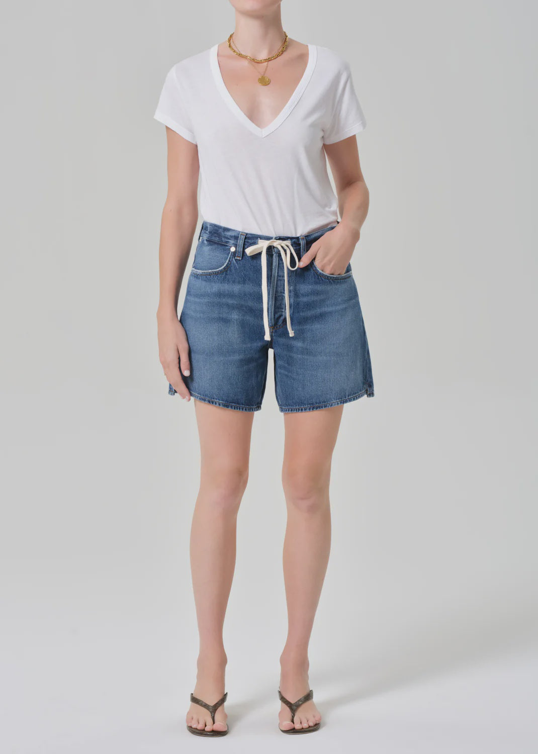 A person wearing Citizens of Humanity/AGOLDE brynn drawstring short in avant (lightwt vintage), a white V-neck T-shirt, flip-flops, a white belt, and layered necklaces stands against a plain light background.