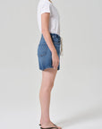 A woman stands sideways against a plain light background, wearing Citizens Of Humanity Brynn drawstring shorts in avant (lightwt vintage), a white t-shirt, and black flip-flops. Her hair is up and arms are relaxed by her side.