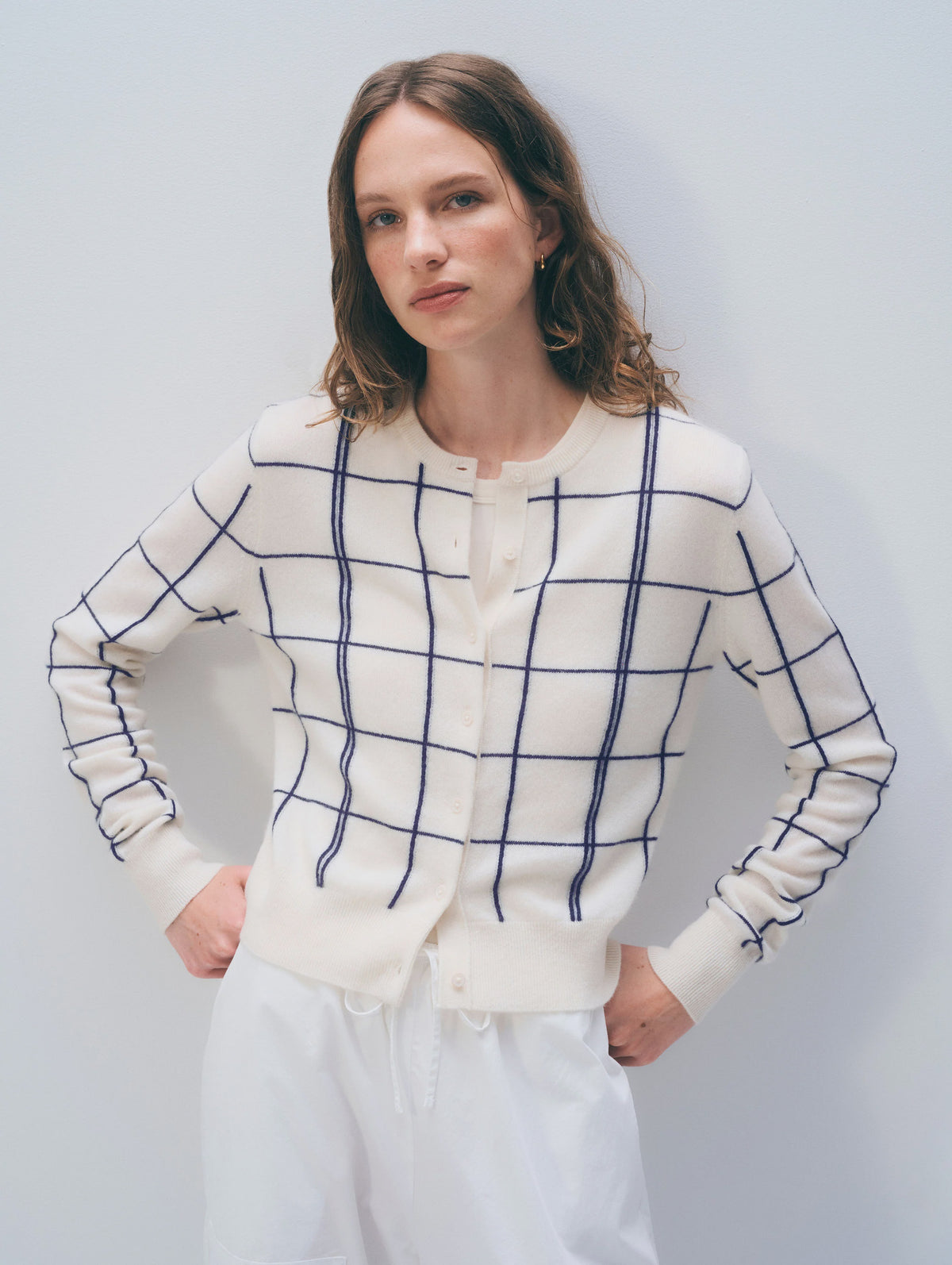 A woman with wavy brown hair wears the White + Warren Cashmere Windowpane Cardigan and white pants, standing against a plain light background with her hands on her hips.