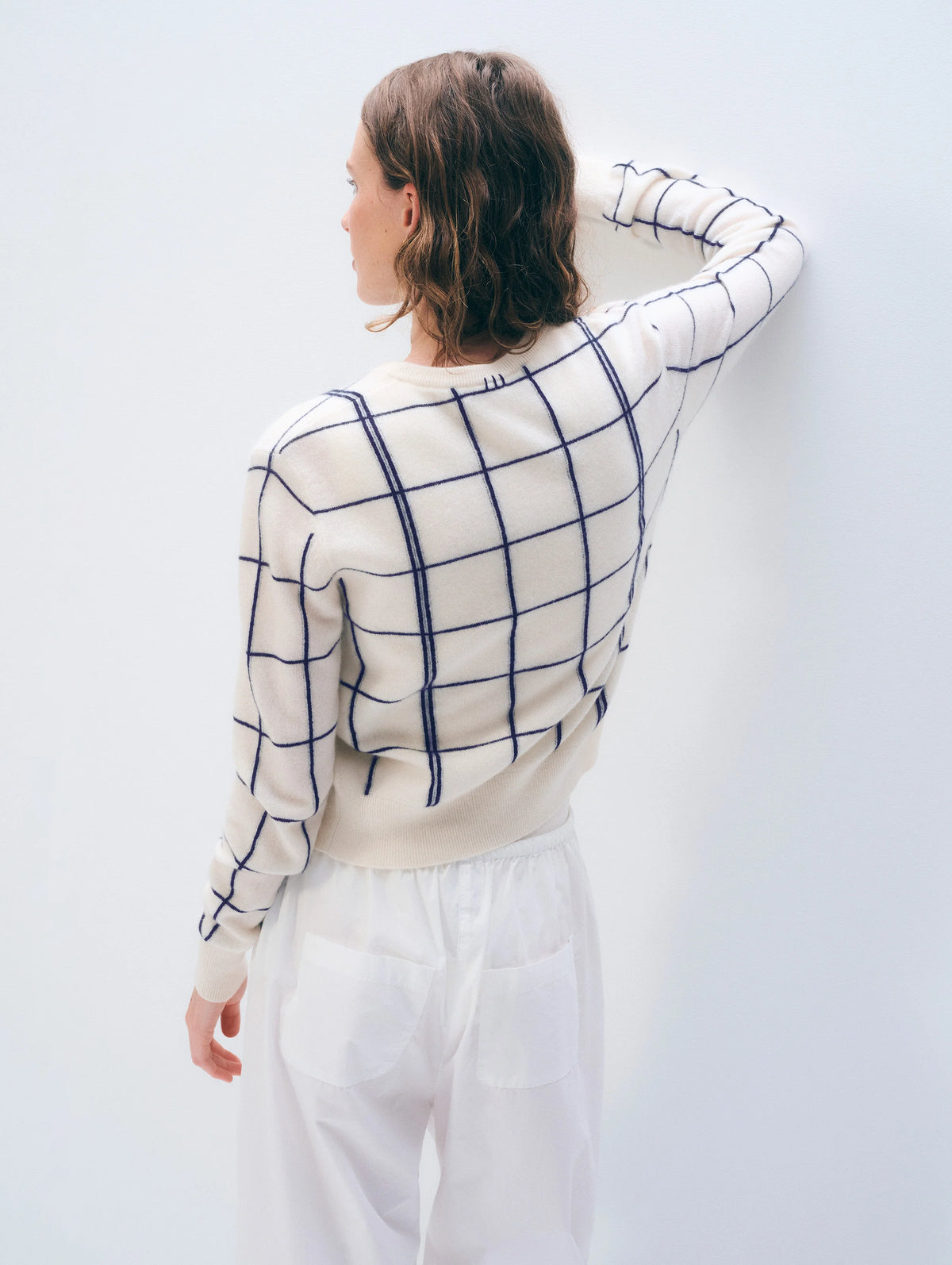 A person with short brown hair faces a white wall, wearing the White + Warren Cashmere Windowpane Cardigan and straight-fit white pants with large back pockets. One arm is bent, touching the wall.