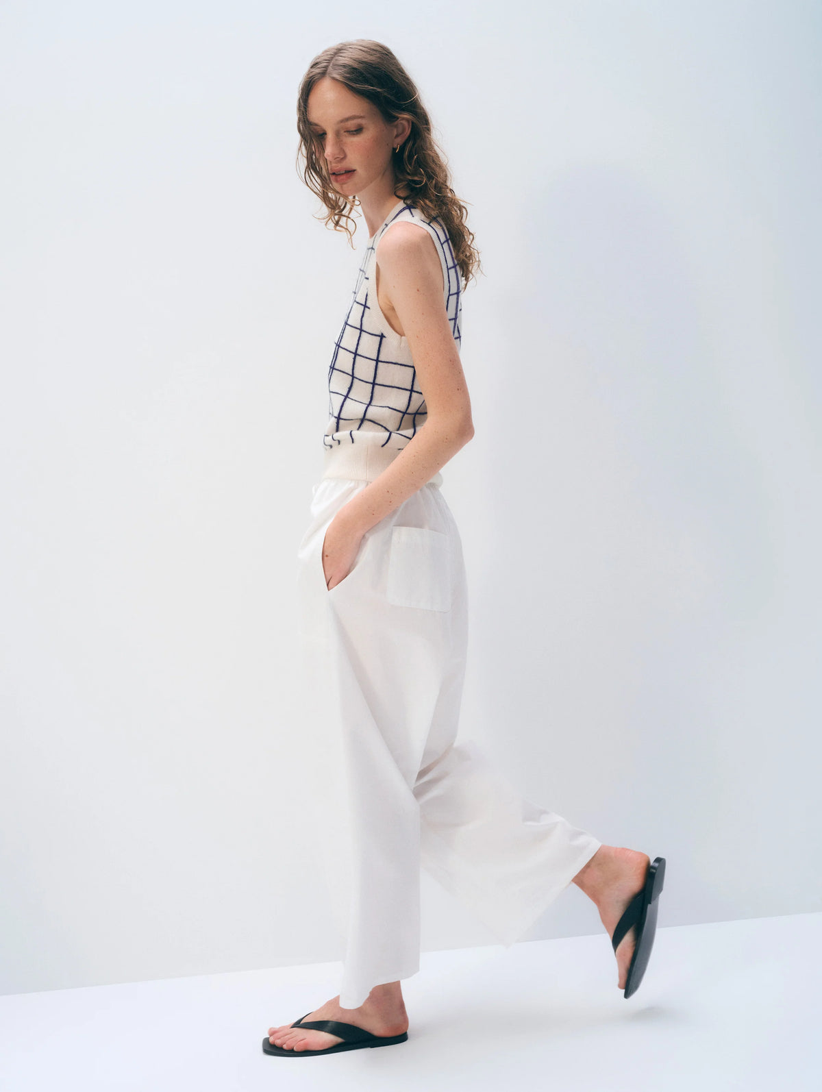 A woman with wavy hair stands sideways against a plain background, wearing the White + Warren Cashmere Windowpane Vest, white high-waisted pocket pants, and black sandals. Her hands are in her pockets as she looks slightly down.