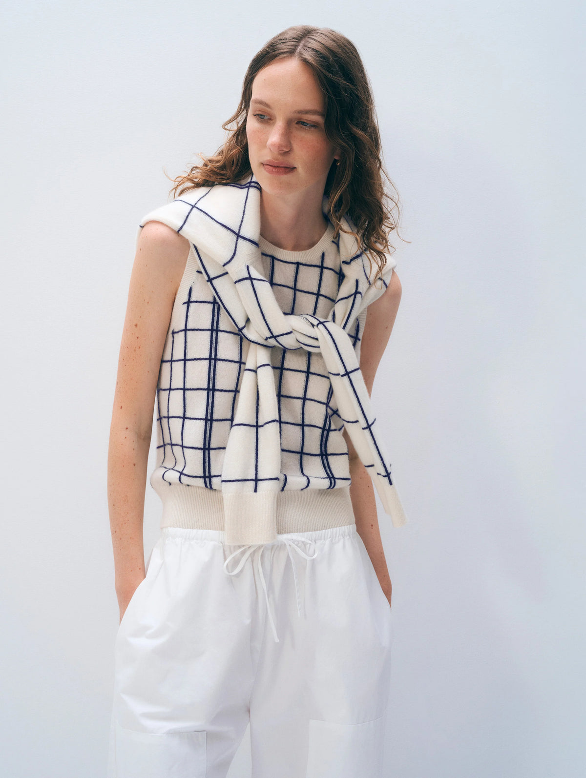 A woman with wavy brown hair wears the White + Warren Cashmere Windowpane Vest and a matching sweater over her shoulders, paired with white pants, standing against a plain light background.