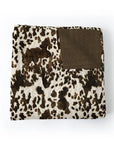 The Faire Bella Hide Brown Quilt features a folded design with a brown and white cowhide pattern and a solid brown corner patch, blending contemporary style with Texas-inspired charm for any space.