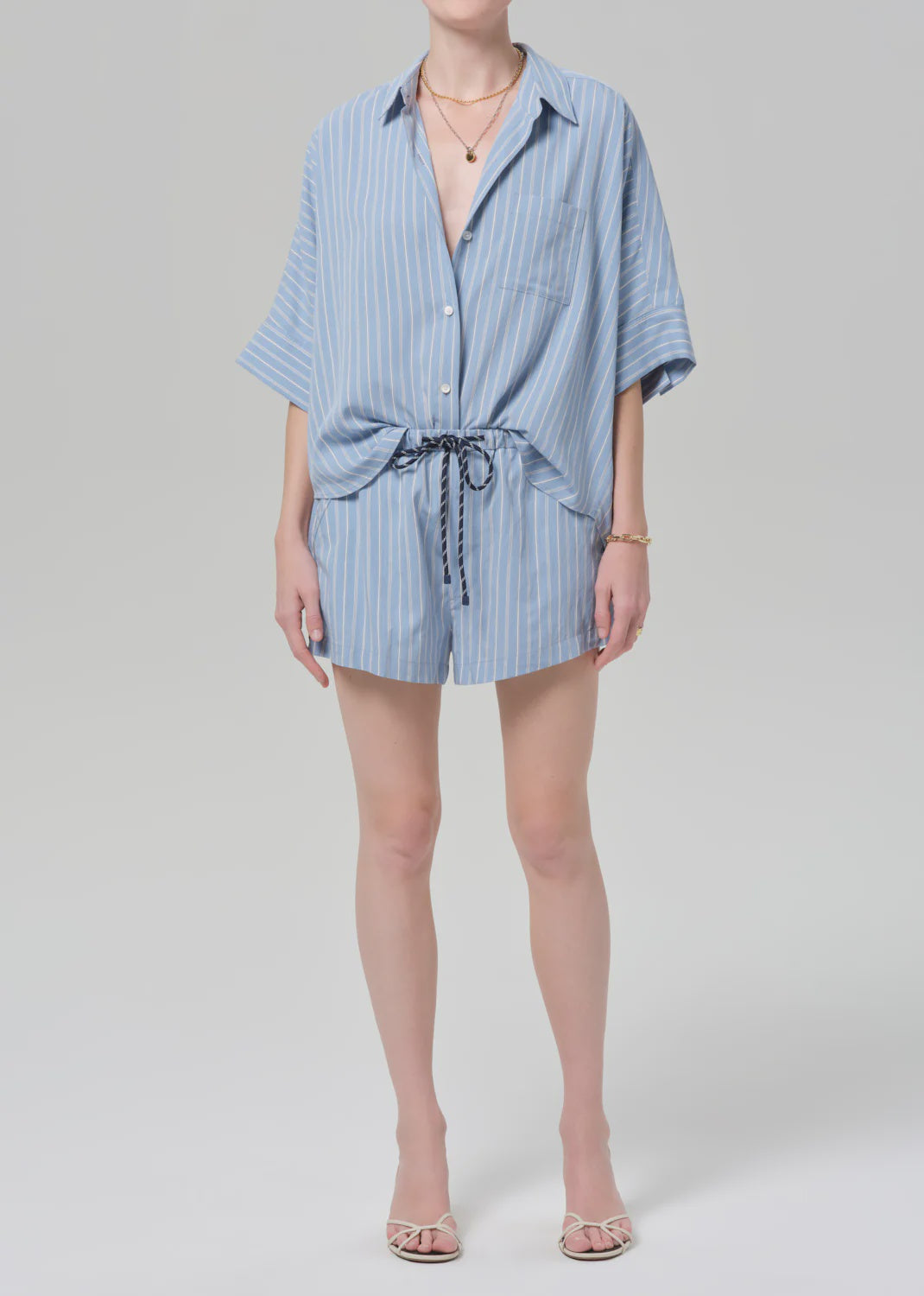 A person wearing Citizens Of Humanity/AGOLDE's este drawstring short in capri stripe, paired with a light blue and white striped button-up, open-toe sandals, and accessorized with a gold necklace and bracelet.