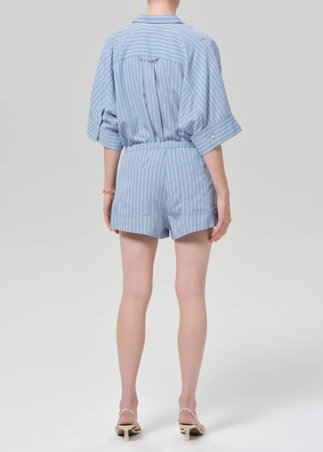 A person with light skin and short hair wears Citizens Of Humanity/AGOLDE's este drawstring short in capri stripe, styled with a matching vertical striped light blue shirt and white low-heel sandals, standing against a plain light gray background.