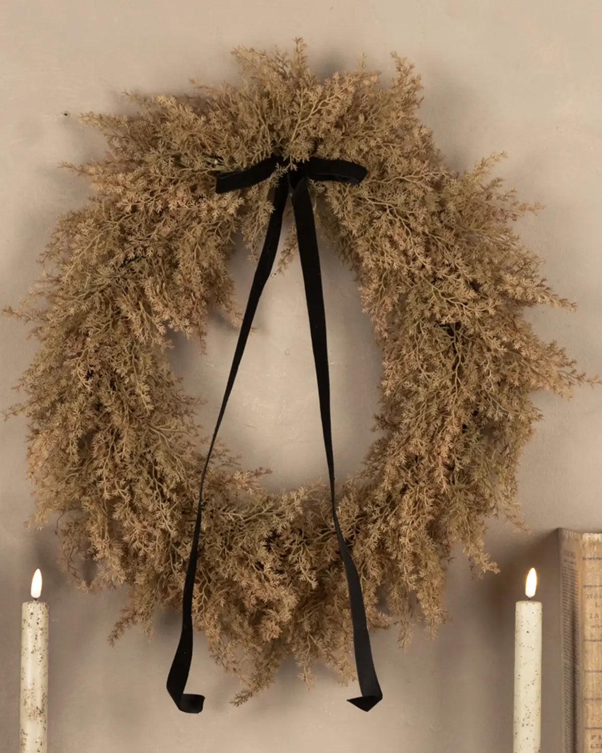 Tan Monterey Cypress wreath with black velvet bow by The Faire displayed above white candles on a beige wall.