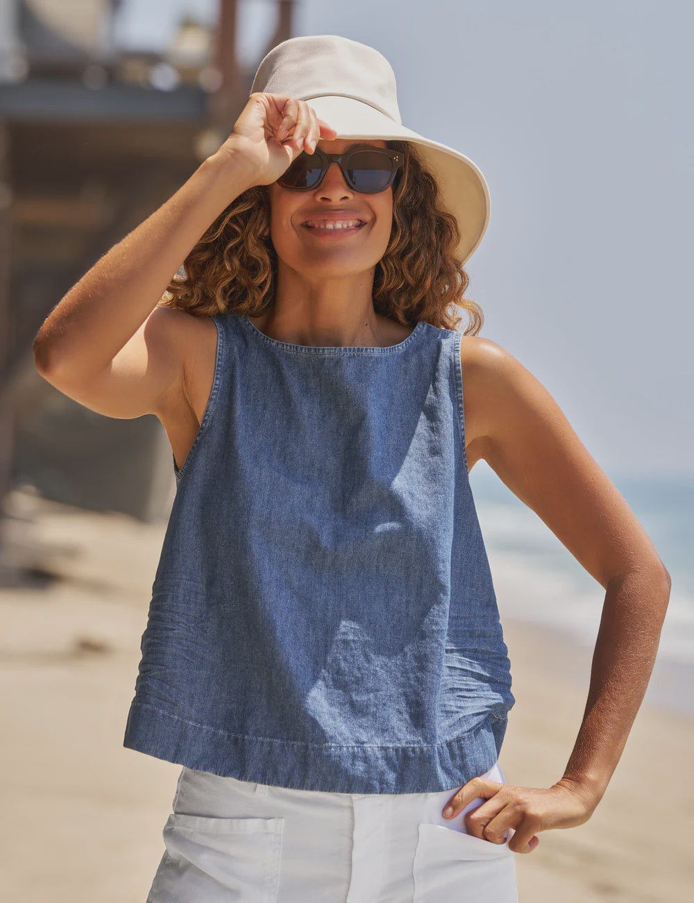 A woman smiles on a sunny beach, wearing a light sun hat, dark sunglasses, and the Frank & Eileen Shell Tank with white shorts. She holds her hat brim as the ocean shimmers in the background.